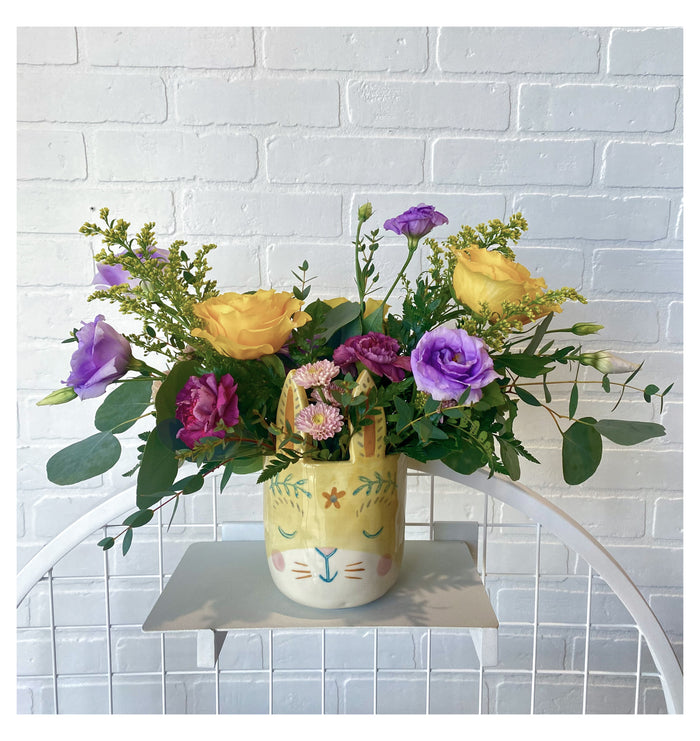 An Easter arrangement in a bunny ceraminc vase with yellow, purple and light purple flowers.